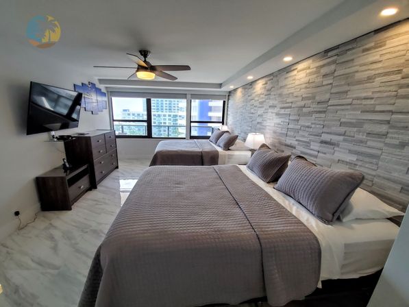 Spacious modern high-rise bedroom with two queen beds, gray quilted bedding, textured stone accent wall, marble floor, ceiling fan, wall-mounted TV and city skyline view through large windows.