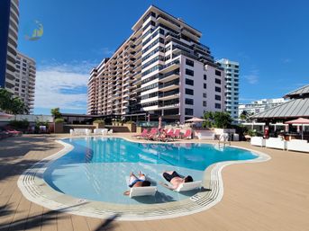 Sunny resort pool and deck with two people relaxing on in-pool loungers, pink chaise lounges and coastal high-rise condos under a bright blue sky.