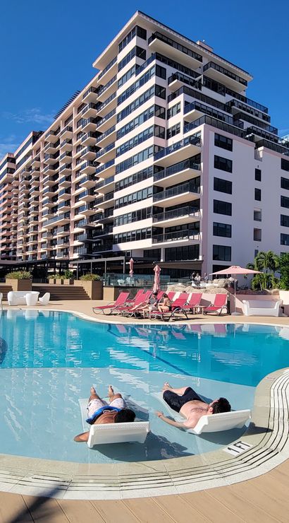 Sunny resort pool and deck with two people relaxing on in-pool loungers, pink chaise lounges and coastal high-rise condos under a bright blue sky.