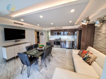 Sleek open-plan downtown condo interior with glossy marble floors, glass dining table and black leather chairs, stainless steel kitchen appliances, wall-mounted TV, recessed lighting, and a white leather sofa with colorful pillows.