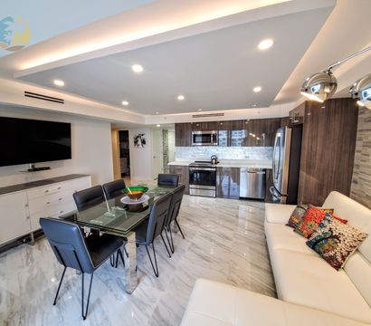 Sleek open-plan downtown condo interior with glossy marble floors, glass dining table and black leather chairs, stainless steel kitchen appliances, wall-mounted TV, recessed lighting, and a white leather sofa with colorful pillows.