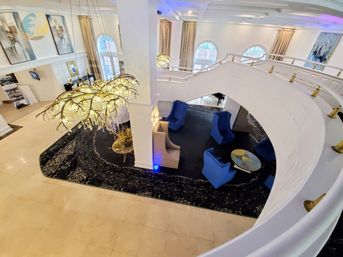 Bird's-eye view of a luxury hotel lobby with a curved white mezzanine railing, oversized branch-like crystal chandelier, black patterned rug seating area with blue armchairs and a round table, beige marble floors and arched windows.