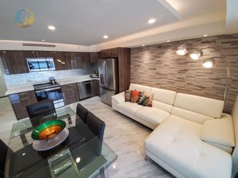 Modern open-plan condo interior with white leather sectional, colorful throw pillows, glass dining table, stainless-steel kitchen appliances, wood-grain cabinets, marble-look floor and textured gray accent wall with arc floor lamp.