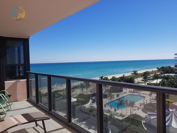 Sunny oceanfront balcony with lounge chairs and glass railing overlooking turquoise sea, sandy beach, palm trees and a resort-style swimming pool under a clear blue sky