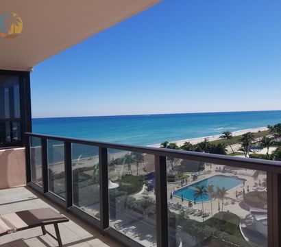 Sunny oceanfront balcony with lounge chairs and glass railing overlooking turquoise sea, sandy beach, palm trees and a resort-style swimming pool under a clear blue sky