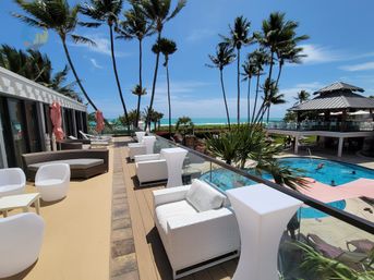 Sunlit tropical oceanfront terrace with white wicker lounge chairs, glass railing, tall palm trees, swimming pool and turquoise sea view