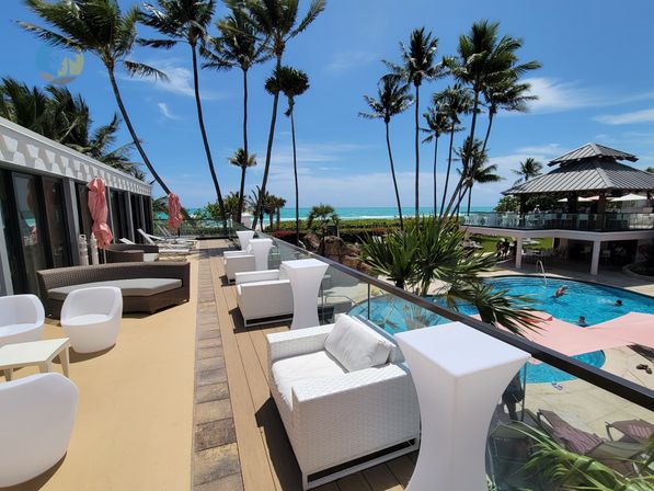Sunlit tropical oceanfront terrace with white wicker lounge chairs, glass railing, tall palm trees, swimming pool and turquoise sea view