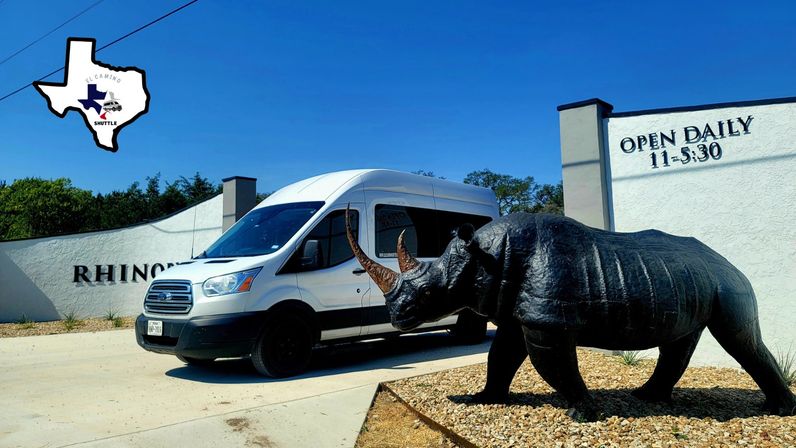 White passenger shuttle van parked beside a stucco building with an "Open Daily 11–5:30" sign, next to a life-size black rhino sculpture and a small Texas-shaped logo against a clear blue sky.
