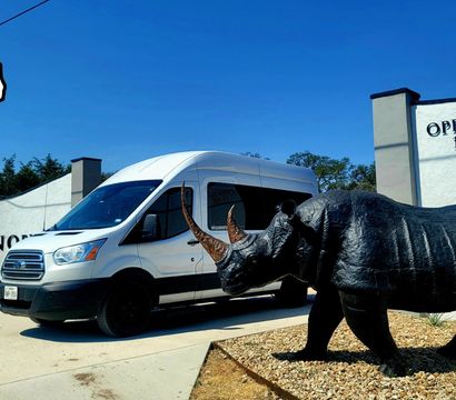 White passenger shuttle van parked beside a stucco building with an "Open Daily 11–5:30" sign, next to a life-size black rhino sculpture and a small Texas-shaped logo against a clear blue sky.