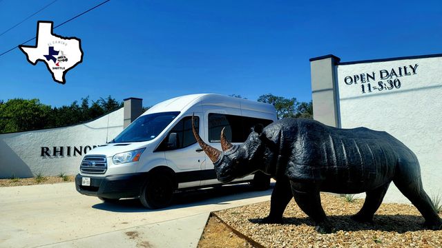 White passenger shuttle van parked beside a stucco building with an "Open Daily 11–5:30" sign, next to a life-size black rhino sculpture and a small Texas-shaped logo against a clear blue sky.
