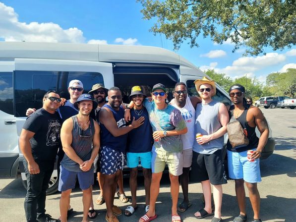 Smiling group of friends in summer clothes and sunglasses posing with canned drinks in front of a white passenger van in a sunny parking lot with trees and blue sky.