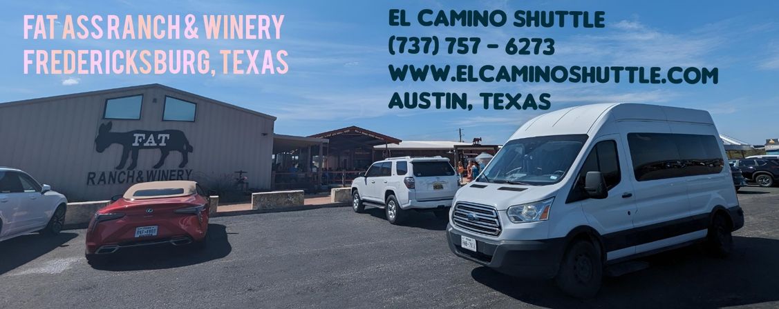 Rustic winery parking lot in Fredericksburg, Texas — barn-style tasting room, parked cars and a white shuttle van under a bright blue sky.