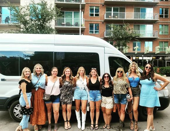 Ten women in summer outfits posing in front of a white passenger van on a downtown city street with brick apartment balconies — playful bachelorette party vibe.
