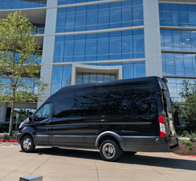 Sleek black high-roof cargo van parked at a curb outside a modern glass office building, trees and blue sky reflected in the windows.