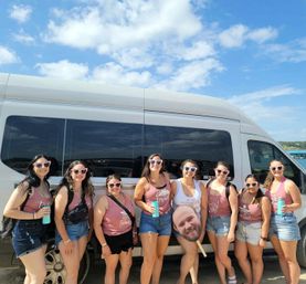 Eight women in matching mauve tank tops and heart-shaped sunglasses posing with drinks and a novelty face cutout in front of a white passenger van at a sunny waterfront — fun girls’ trip/bachelorette vibe.