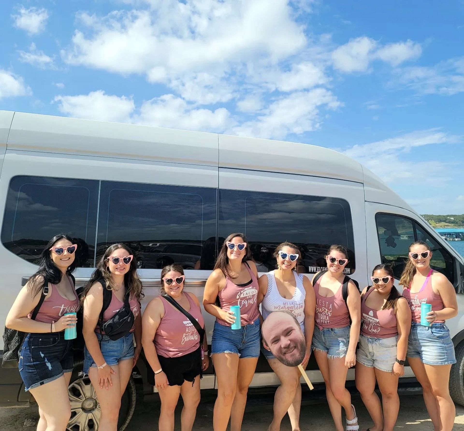 Eight women in matching mauve tank tops and heart-shaped sunglasses posing with drinks and a novelty face cutout in front of a white passenger van at a sunny waterfront — fun girls’ trip/bachelorette vibe.