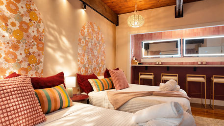 Cozy boho guest room with twin beds, retro orange floral arch murals, textured pink and striped pillows, warm pendant light, LED-lit vanity with stools and exposed wooden beam ceiling