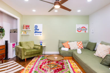 Cozy colorful living room with a green velvet armchair and olive sectional sofa, bright multicolor shag rug, round wooden coffee table, pastel green accent wall, ceiling fan and recessed lights.