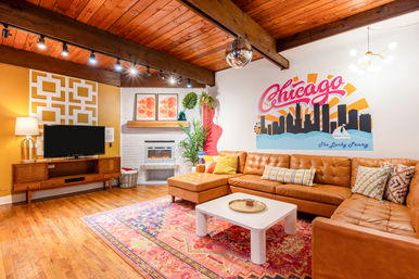 Bright midcentury-modern living room with a tan leather sectional, colorful area rug, white coffee table, wood‑beamed ceiling with disco ball, fireplace, TV console, plants, and a large Chicago skyline mural