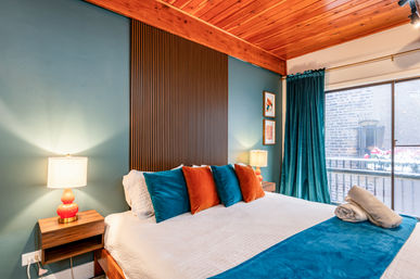 Cozy modern urban apartment bedroom with wooden ceiling and slatted headboard, teal accent wall and curtain, white bed with teal and orange pillows, bedside lamps and sliding glass balcony overlooking a brick building