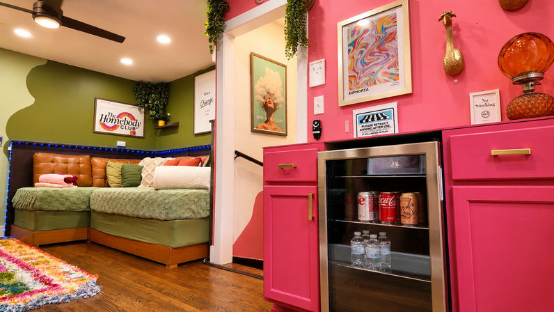 Cozy colorful lounge with hot-pink cabinets and mini fridge, green accent wall, sectional daybed with throw pillows, framed art, hanging plant, LED strip lights and hardwood floor
