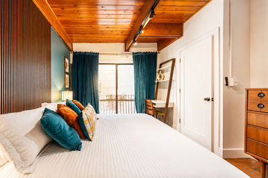 Cozy modern urban bedroom with exposed wood-beamed ceiling, slatted wood accent wall, white quilted bed, teal and rust throw pillows, teal curtains over sliding balcony door, ladder desk and mid-century wooden dresser