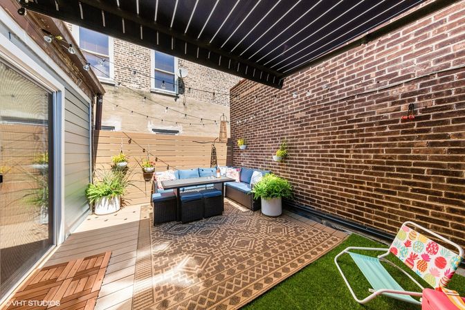 Cozy urban patio with exposed brick walls, black slatted canopy, L-shaped blue-cushioned sectional, patterned outdoor rug, string lights, potted greenery and a colorful lawn chair.