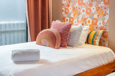 Cozy retro-style bedroom with white quilted bed and rolled towel, velvet peach curtains, colorful decorative pillows and orange floral wallpaper