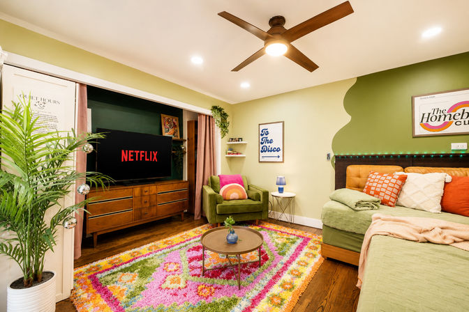 Colorful cozy living room with TV showing a streaming screen, green velvet armchair with rainbow cushion, bright floral shag rug, mid-century wood console, tufted daybed with patterned pillows, ceiling fan and potted plant