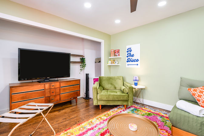 Bright cozy living room with a large flat-screen TV on a mid-century wood console, olive-green armchair, round coffee table, colorful shag rug, pastel green walls and playful "To The Disco" wall art.