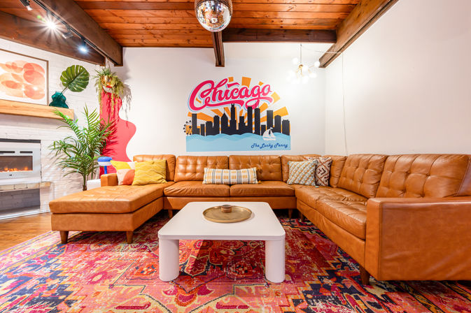 Bright Chicago-themed lounge with caramel leather sectional, vibrant patterned rug, white coffee table, indoor plants, fireplace, disco ball and Chicago skyline mural under exposed wooden beams.