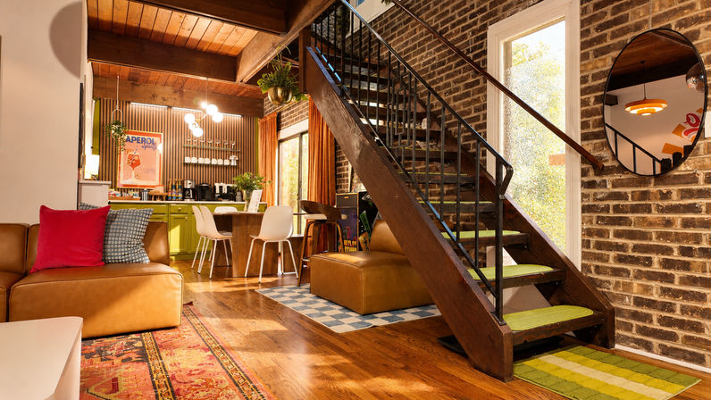 Cozy mid-century modern urban bungalow interior with a leather sofa and colorful pillows, wooden staircase against an exposed brick wall, green open kitchen cabinets, pendant lights and hanging plants.