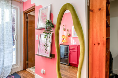 Bright pink home entryway with hardwood floor, a green oval full-length mirror reflecting a small pink kitchenette with gold accents, floating wall shelves with framed art and a trailing plant, and a sliding glass door with patterned curtain.