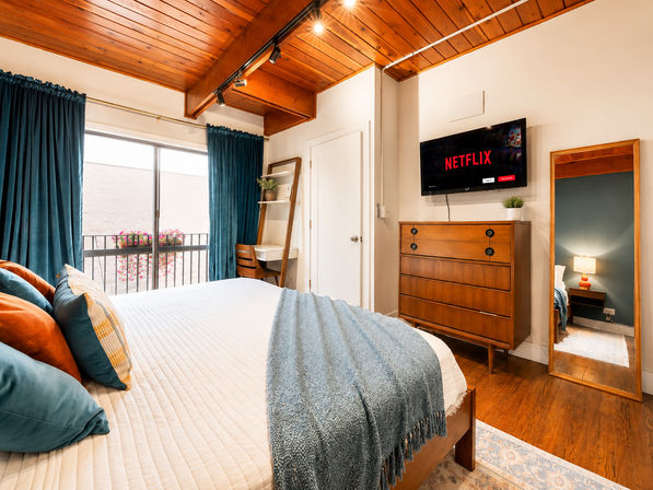 Cozy bright bedroom with wooden vaulted ceiling and mid-century furniture — queen bed with white quilt and teal throw, teal curtains opening to a balcony with flower boxes, wall-mounted TV showing Netflix, full-length mirror and small desk.