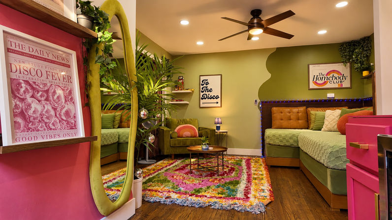 Cozy retro-styled living room with olive-green accent wall, velvet green armchair and matching daybed, colorful shag rug, round coffee table, tall oval mirror, tropical potted plant, disco-themed posters and string lights.