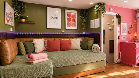 Vibrant cozy living room corner with olive-green walls and pink accent, quilted green daybed with leather back cushions, coral and cream throw pillows, folded pink towels, hanging plants, framed art, purple LED strip lighting and disco-ball pendants.