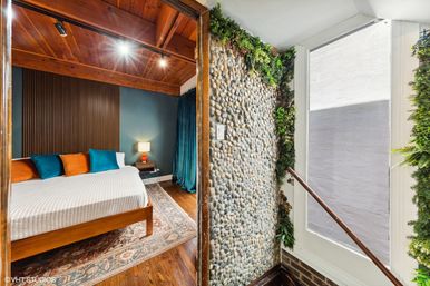 Cozy bedroom with wooden vaulted ceiling, mid-century wood bed dressed in white linen and teal and orange pillows, dark blue accent wall with nightstand lamp, and an adjacent pebble stone accent wall with vertical greenery beside a bright stairwell window.
