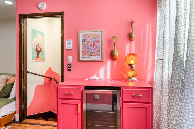 Vibrant pink interior nook with coral cabinets and mini fridge, amber retro lamp, abstract wall art, gold gourd wall decor, patterned curtains and an open doorway to a stairwell.