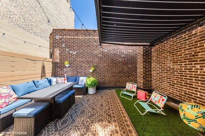 Urban rooftop patio with brick walls and string lights; wicker sectional with blue cushions and long dining table on a patterned rug, hanging planters and potted fern, artificial turf area with colorful low beach chairs and a cooler under a retractable awning.