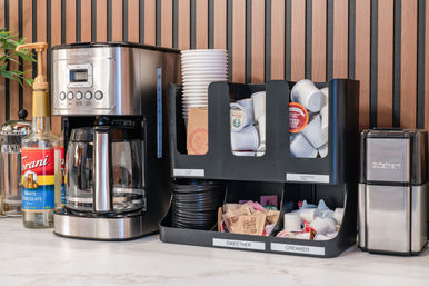 Office coffee station on a marble countertop with a stainless-steel drip coffee maker, syrup bottles, stacked disposable cups, single-serve coffee pods, and labeled sweetener and creamer organizers against a modern wood-slat wall.