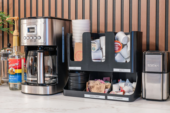 Office coffee station on a marble countertop with a stainless-steel drip coffee maker, syrup bottles, stacked disposable cups, single-serve coffee pods, and labeled sweetener and creamer organizers against a modern wood-slat wall.