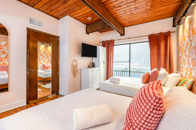 Bright cozy guest bedroom with exposed wooden ceiling beams, two beds with white quilts and coral textured pillows, floral accent wall, flat-screen TV on a white dresser and sliding glass door with coral curtains to a balcony.