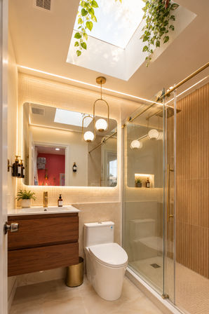Bright modern bathroom with skylight and hanging plants, backlit mirror and globe pendant lights, glass shower with gold hardware, wood floating vanity, white toilet and warm beige tile