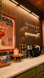 Cozy coffee bar with marble countertop and green cabinets, wood‑slat accent wall and warm LED lighting, glass shelves of mugs and glasses, syrup pumps and drip and single‑serve coffee makers, Aperol Spritz poster.
