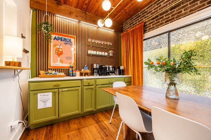 Cheery mid-century modern dining nook with green lower cabinets and marble countertop coffee station, striped wood accent wall with orange cocktail poster, velvet orange curtains, wooden ceiling and table, white chairs, and a vase of citrus branches by a large sunlit window