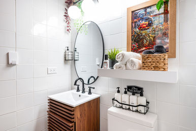 Bright modern small bathroom with glossy white tile, round black-framed mirror above a wooden slatted vanity, black faucet, floating shelf holding rolled towels, wicker basket, plant and framed artwork, with labeled soap dispensers on the toilet — cozy spa-like styling.