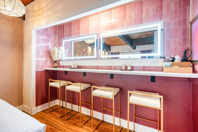 Stylish pink-tiled vanity/dressing bar with LED-lit rectangular mirror, white countertop, pampas grass in a vase, and four gold-frame stools with white cushions on hardwood floors.