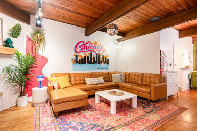 Cozy mid-century modern lounge with tan leather L-shaped sectional on a colorful patterned rug, white coffee table, Chicago skyline mural on the wall, wooden beamed ceiling, hanging plants and a disco ball.