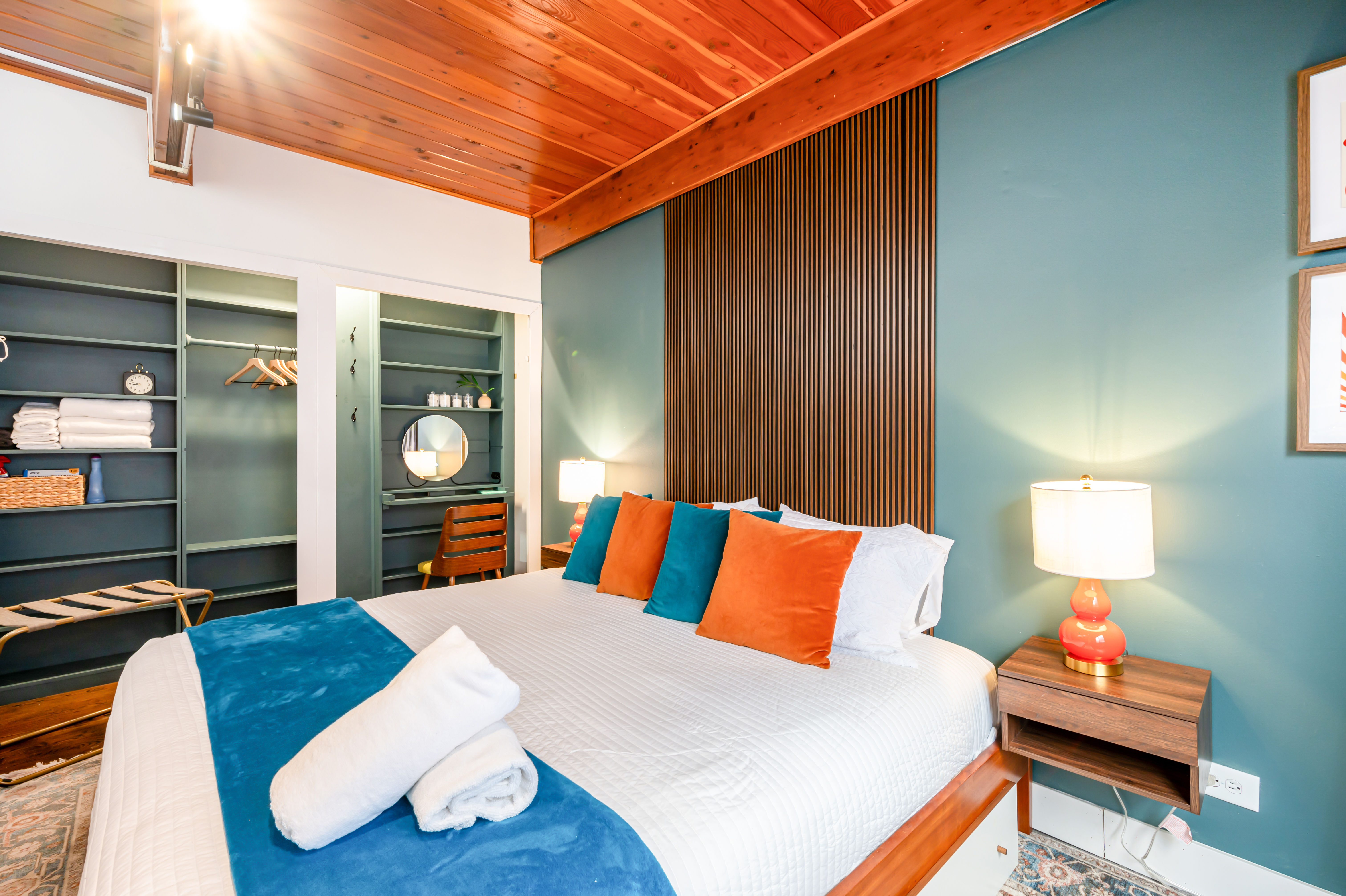 Cozy mid-century modern bedroom with vaulted wood ceiling and teal accent wall, vertical wood-slat headboard, king bed dressed in white linens with teal runner and orange and teal throw pillows, bedside lamp on a wooden nightstand, open shelving closet and small vanity with round mirror.