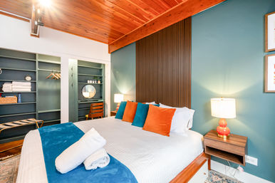 Cozy mid-century modern bedroom with vaulted wood ceiling and teal accent wall, vertical wood-slat headboard, king bed dressed in white linens with teal runner and orange and teal throw pillows, bedside lamp on a wooden nightstand, open shelving closet and small vanity with round mirror.